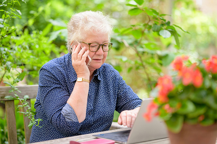 En äldre kvinna sitter vid datorn och pratar i mobiltelefon.